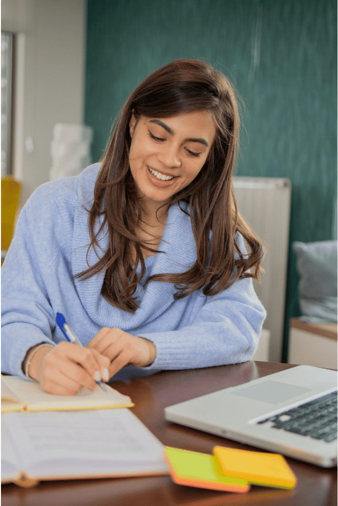 Woman smiling and writing at desk with laptop.